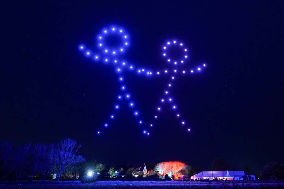 A night sky is illuminated by a mesmerizing drone show forming two stick figures holding hands. Below the light display, tree silhouettes and small buildings are visible. The scene is vibrant with colored lights, giving it a festive feel.