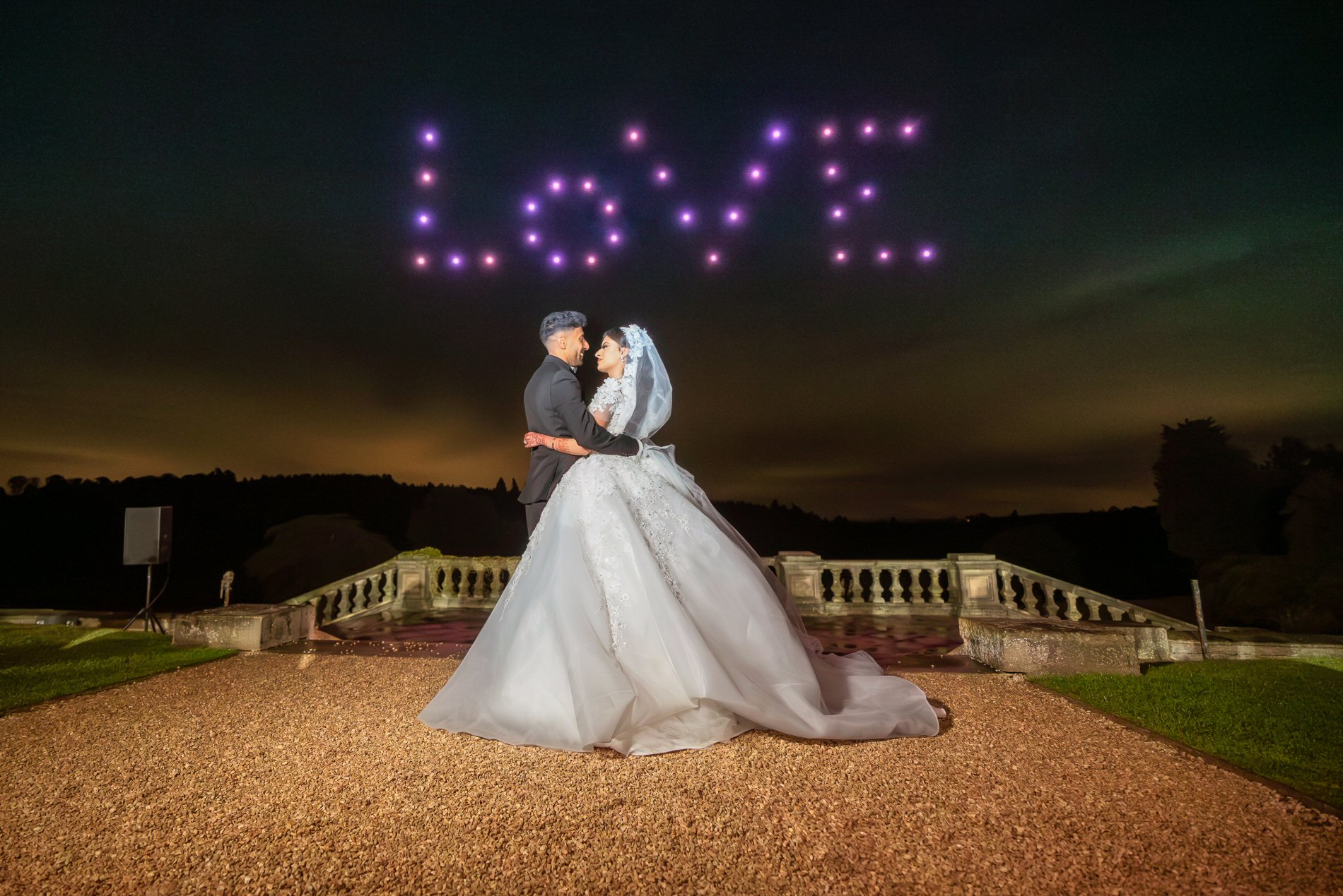 A bride and groom stand facing each other at night, holding hands. The bride wears a white gown and veil, and the groom is in a dark suit. Above them, drones have created the word "LOVE" in the sky with illuminated lights. A scenic outdoor backdrop surrounds them.