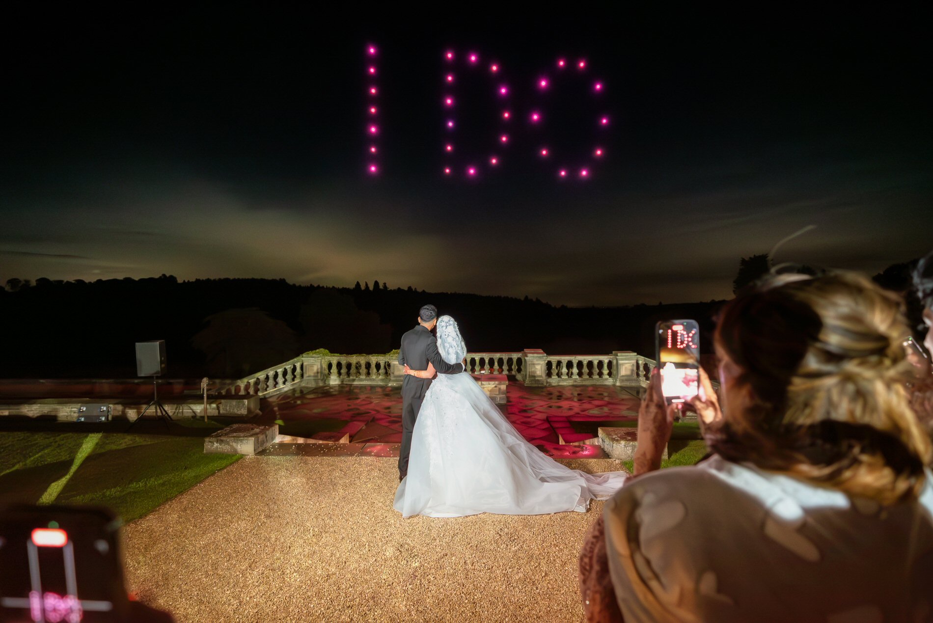 A bride and groom stand on a terrace, looking at the night sky where the words "I DO" are displayed in pink lights. Several people in the foreground capture the moment on their phones.