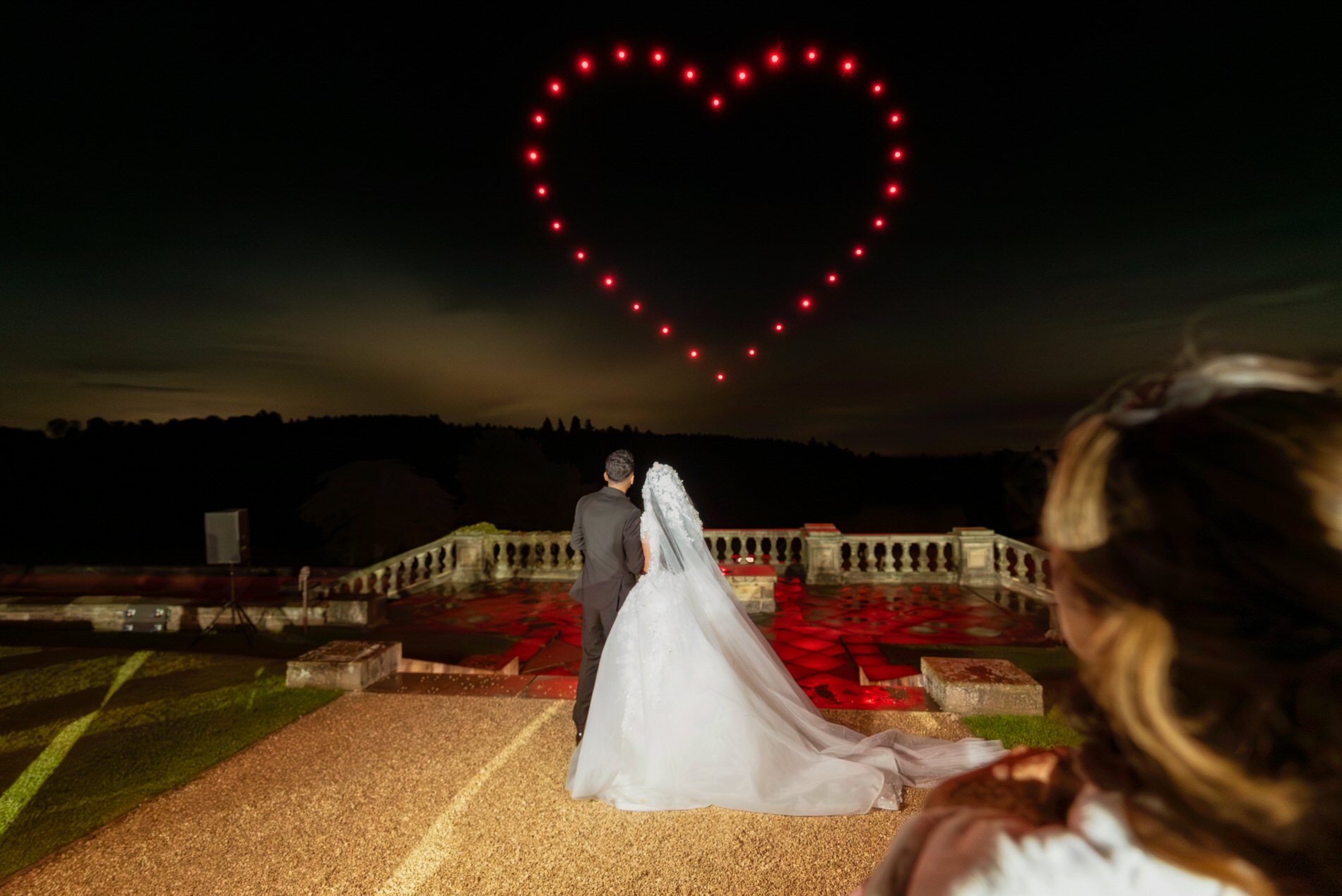 A couple, in wedding attire, stands on a pathway looking at a night sky where lights form a heart shape. An onlooker, partially visible, stands in the foreground on the right. The setting appears to be an outdoor venue with a lit stage and balustrade in the background.