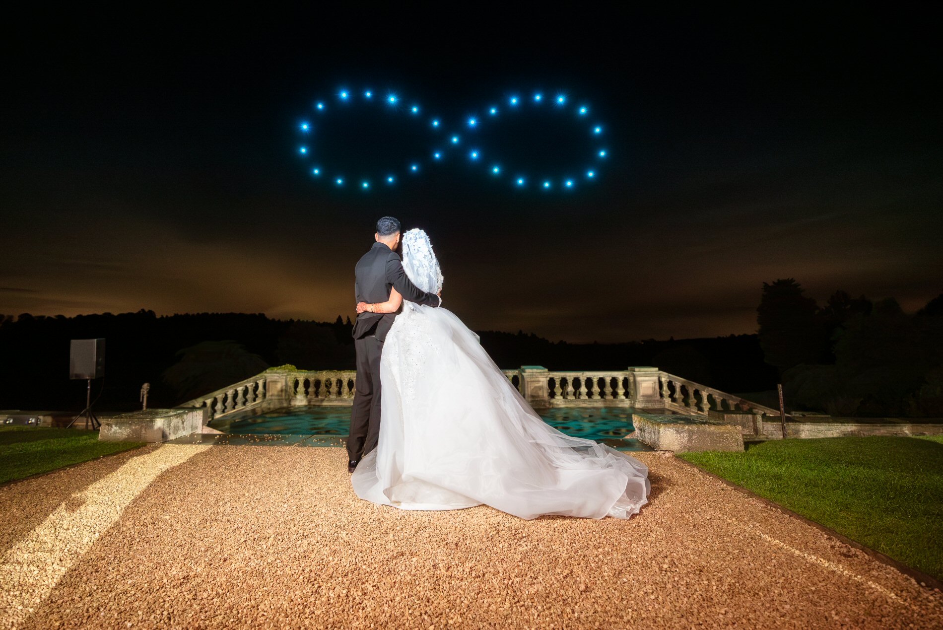 A newlywed couple stands outside at night, looking at a drone show