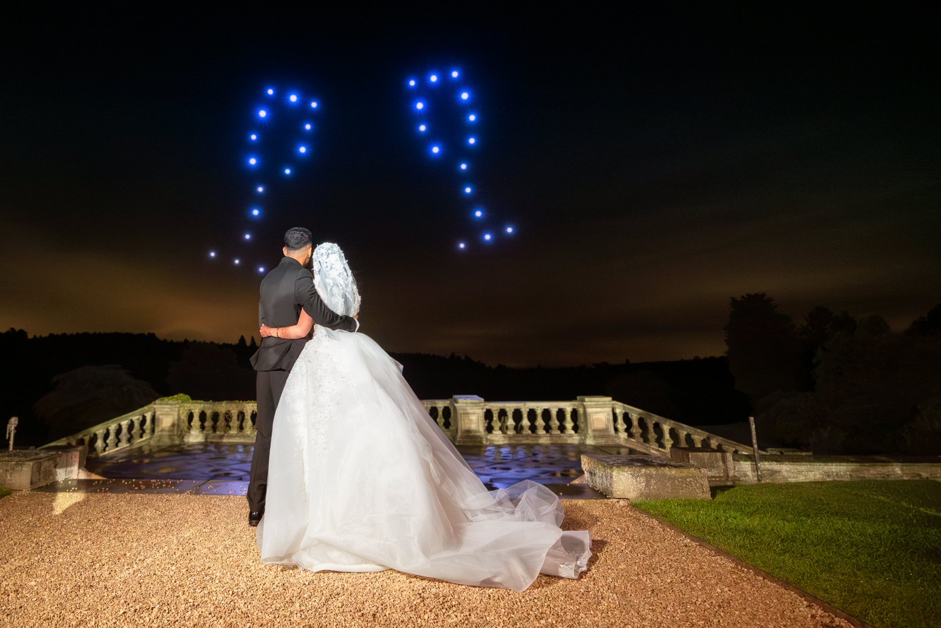 A bride and groom stand facing a nighttime sky, illuminated by the glow of arranged drone lights forming the shape of a heart. They stand on a gravel surface in front of a stone railing, looking out from an elevated terrace.