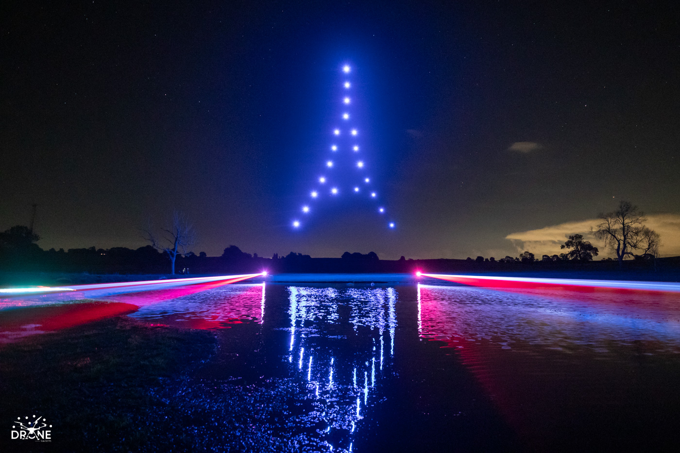 A nighttime scene with a drone laser light show creates the outline of the Eiffel Tower in the sky. Brilliant blue lasers form the tower, while streaks of red and blue are reflected on a body of water below, silhouetting trees in the background.