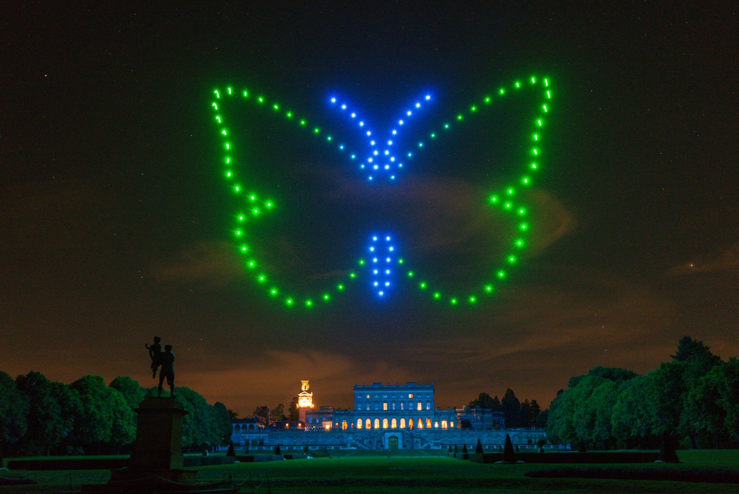 A butterfly shape created with blue and green drones is illuminated in the night sky above a large historic building with trees and a statue in the foreground.