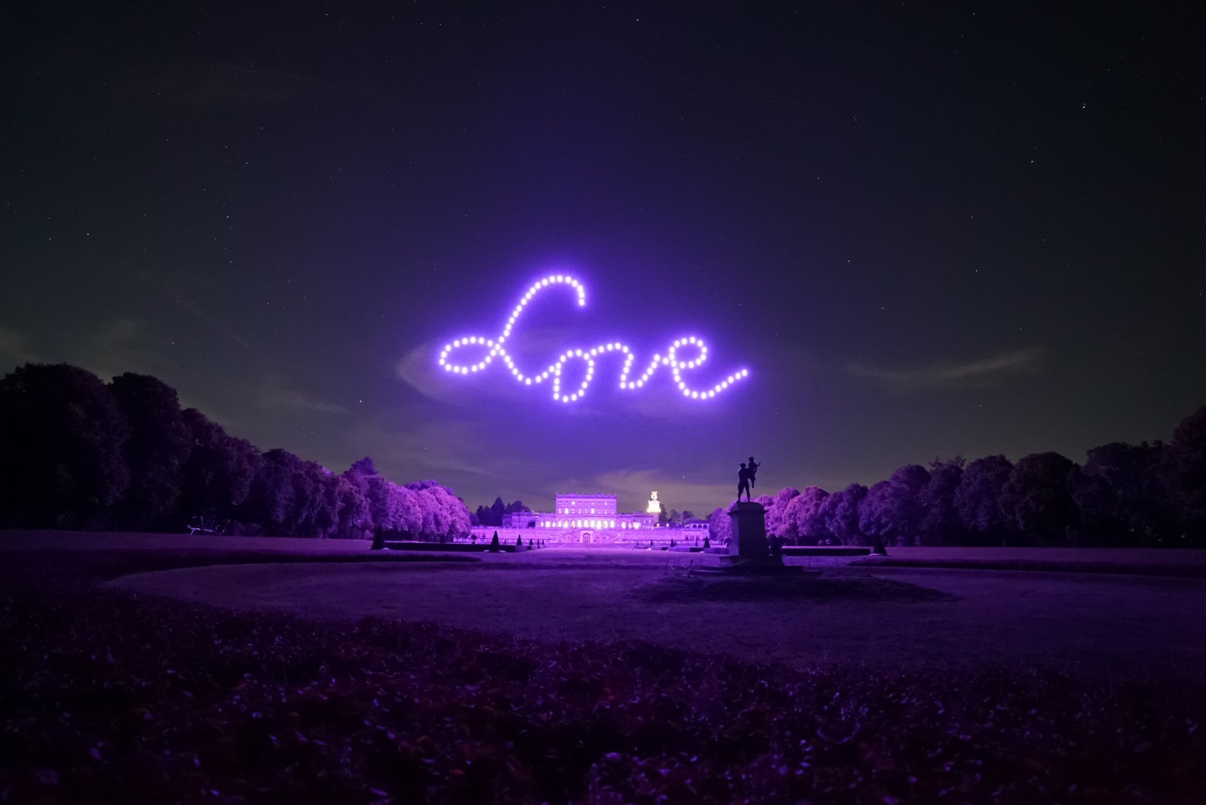 A nighttime scene in a park shows the word "Love" written in the sky with purple lights, likely created by drones. A large building is visible in the background, with trees lining both sides and a statue in the center foreground.