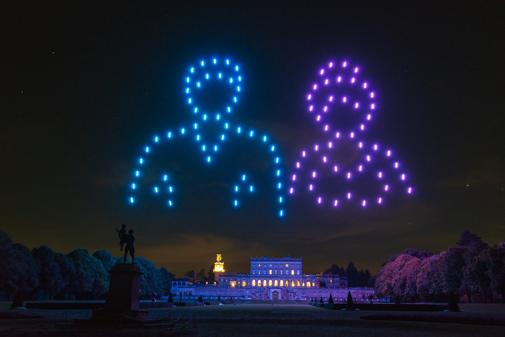 Two large outlines of human figures made from blue and purple lights appear in the night sky above a grand, illuminated building and garden with trees and a statue in the foreground.