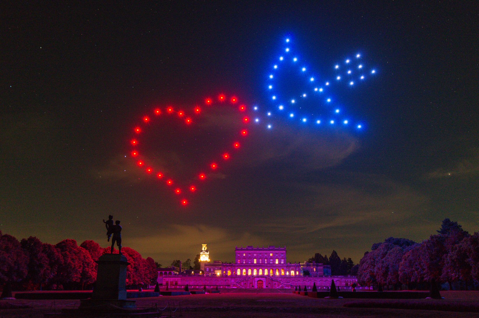 A night scene shows illuminated drones forming a red heart and a blue dove in the sky above a large, well-lit historic building, with a silhouetted statue and trees in the foreground.