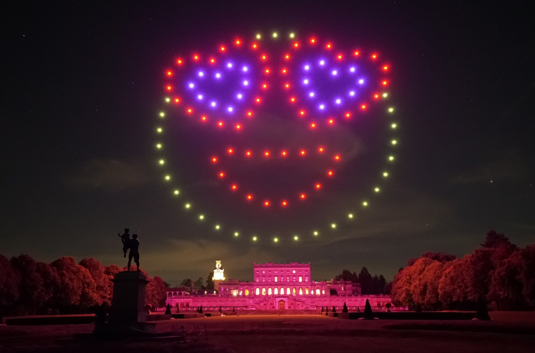 A nighttime scene shows a large smiley face with heart-shaped eyes made of red, yellow, and purple lights formed by drones above an illuminated building and a statue in a park setting.
