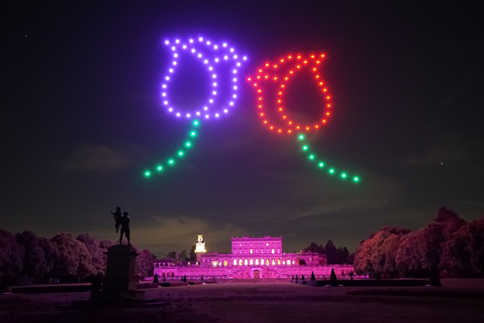 A nighttime scene showing two flower shapes formed by red, purple, and green drones in the sky above a large, illuminated building with a clock tower and statues in the foreground.