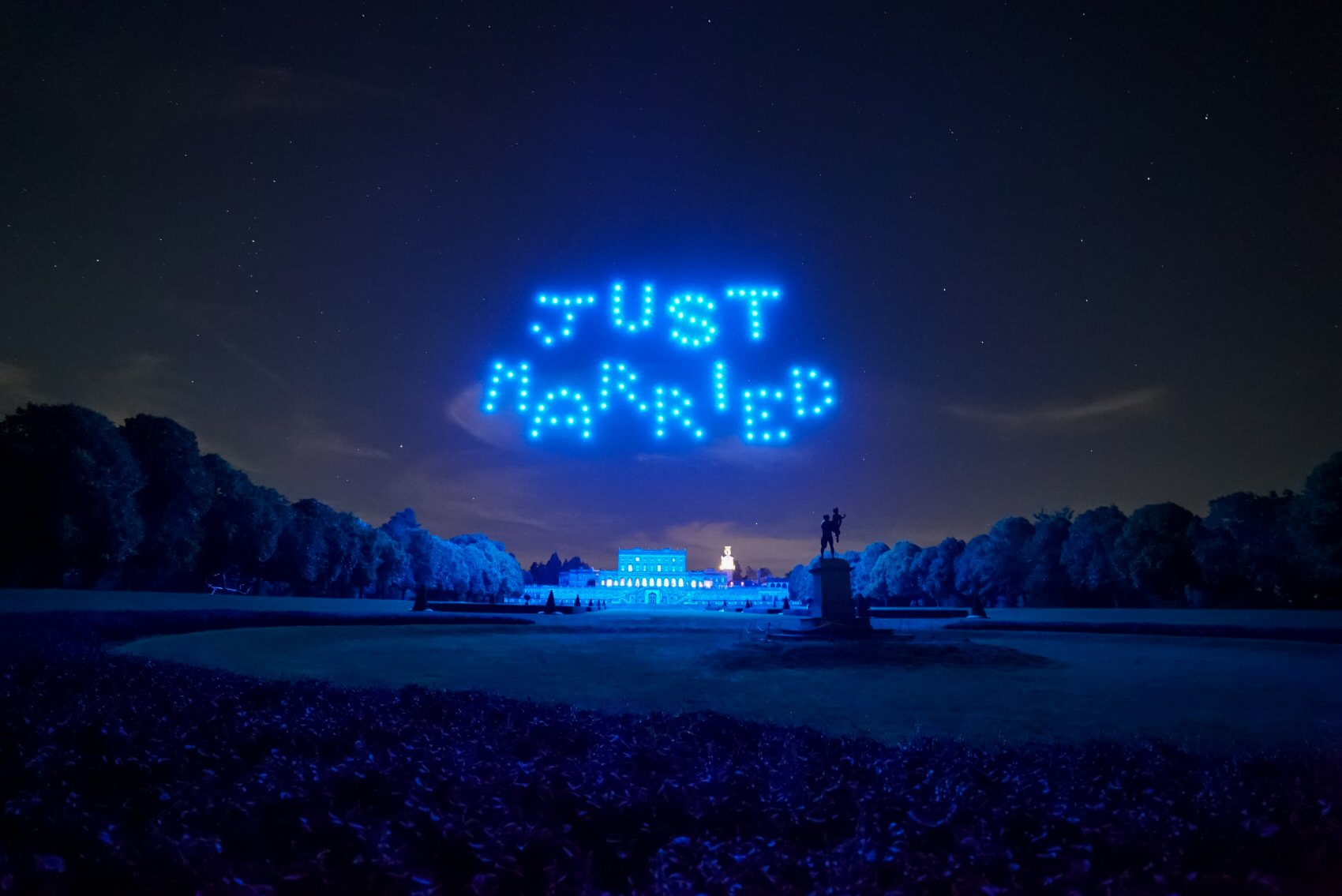 A nighttime scene shows blue drones forming the words "JUST MARRIED" in the sky above a large building and a park with trees and a statue visible in the foreground.