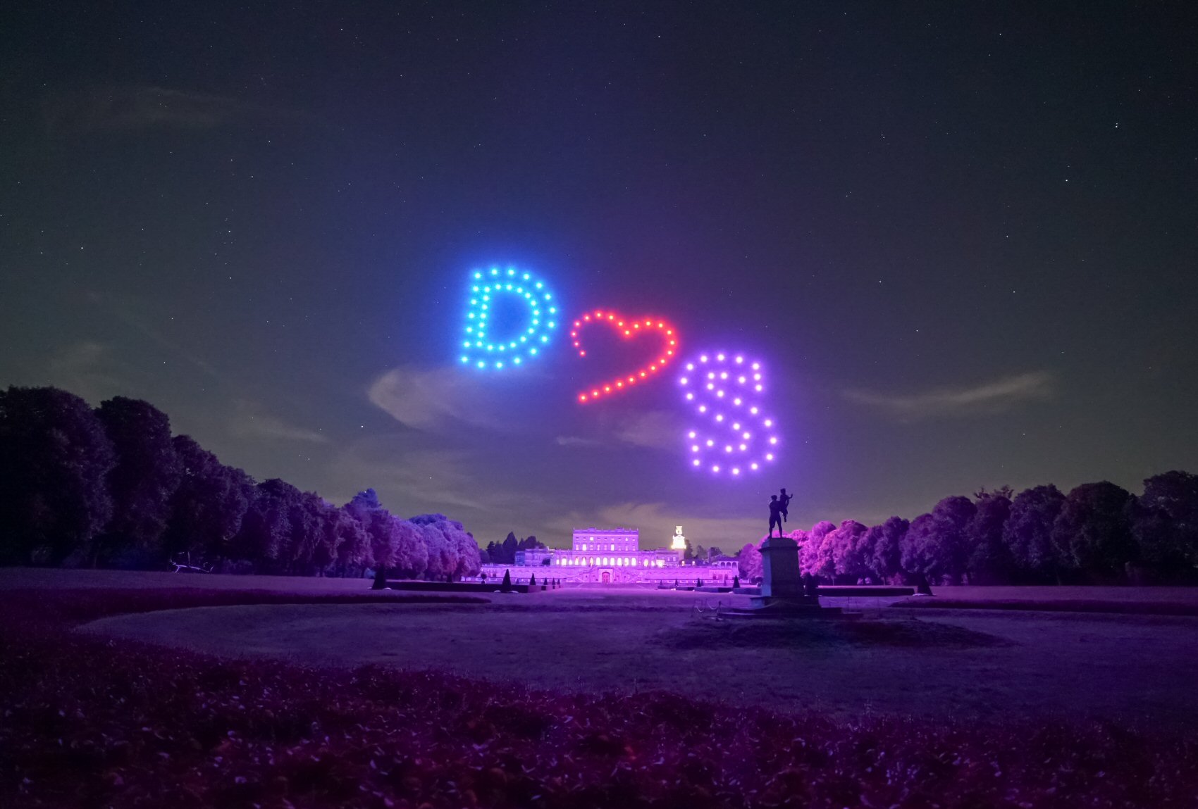 A nighttime scene shows illuminated drones forming the shapes “D ♥ S” in the sky above a large building, with trees and a statue visible in the foreground.