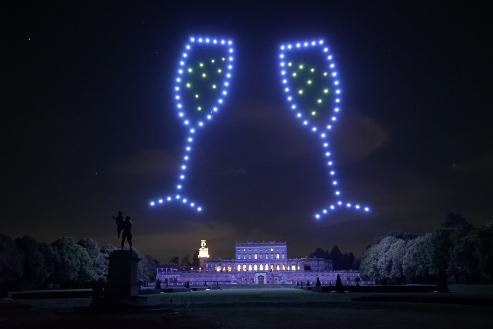 A nighttime scene featuring a large historic building with illuminated windows. Above the building, drones form the shapes of two clinking champagne glasses in the sky. A statue is visible in the foreground.