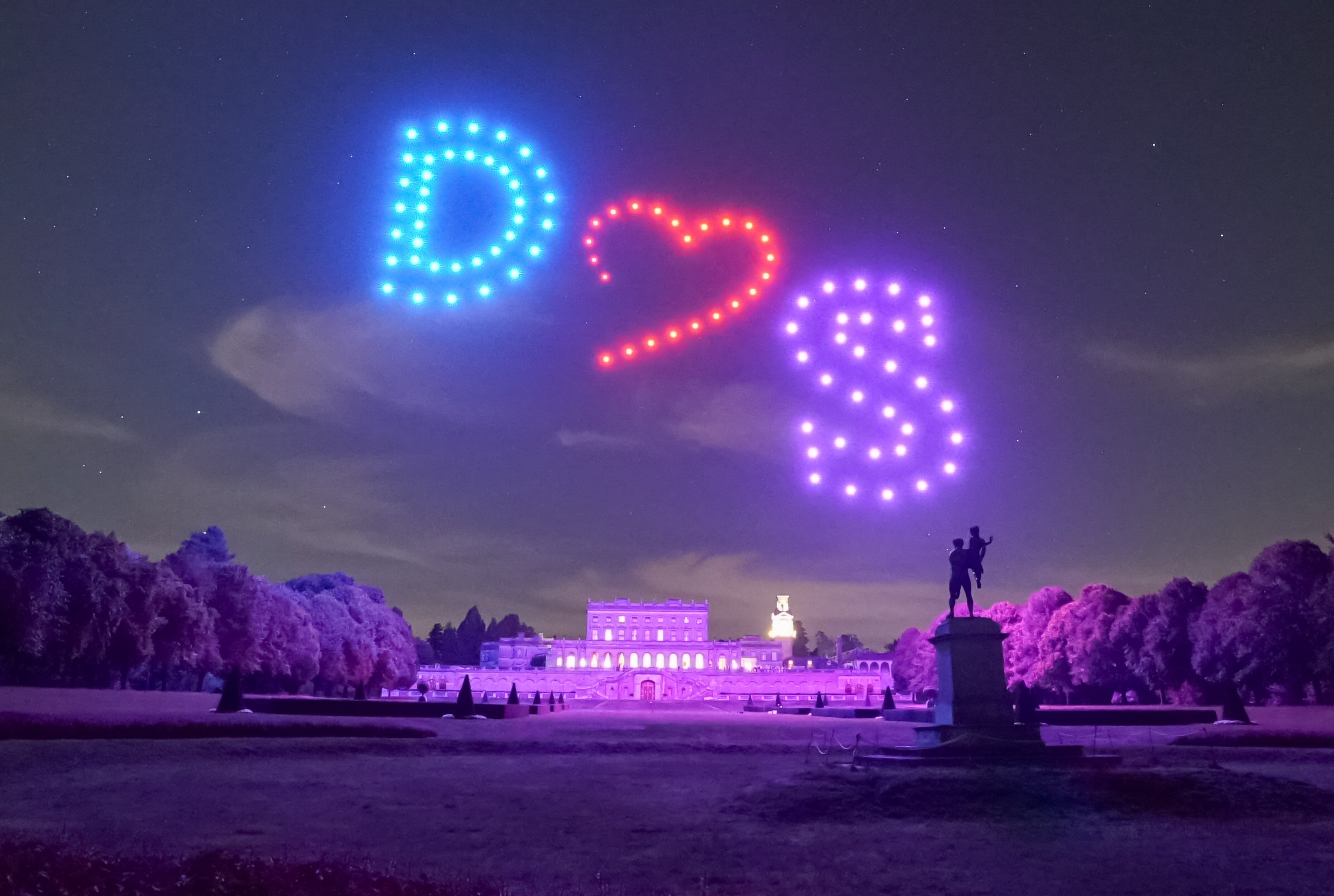 A nighttime scene of an illuminated park with drones in the sky forming the letters "D" and "S" and a heart symbol between them above a large, lit-up building and a statue in the foreground.